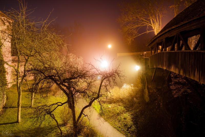Long exposure of an old medieval bridge in Rothenburg ob der Tauber with a strong orange light illuminating a tree at night.